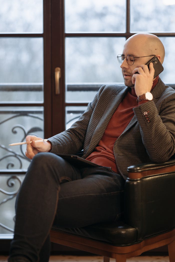 Adult man in casual business attire sitting in an armchair, talking on cellphone, taking notes.